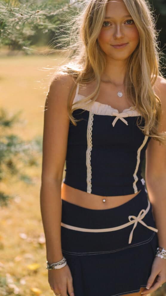 A young woman with long blonde hair stands outdoors in sunlight, wearing a navy blue crop top and matching skirt with cream lace details. She has a navel piercing, bracelets, and a necklace, and is smiling softly. by Justin Salem Meyer