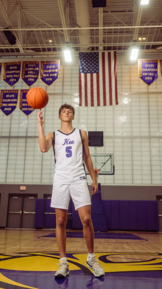 A basketball player in a white Kee uniform with the number 5 spins a basketball on his finger in a gym. Championship banners and an American flag hang on the wall behind him. by Justin Salem Meyer