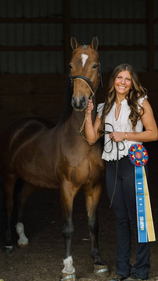 A smiling young woman stands beside a brown horse inside a stable, holding its lead and a large blue and red award ribbon with streamers. The woman is wearing a white blouse and black pants. by Justin Salem Meyer