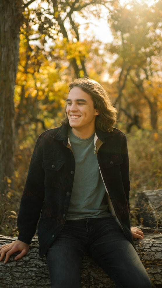 A young person with long hair sits on a log in a sunlit forest, smiling and looking to the side. They wear a green shirt and a dark jacket, surrounded by autumn trees with golden leaves. by Justin Salem Meyer