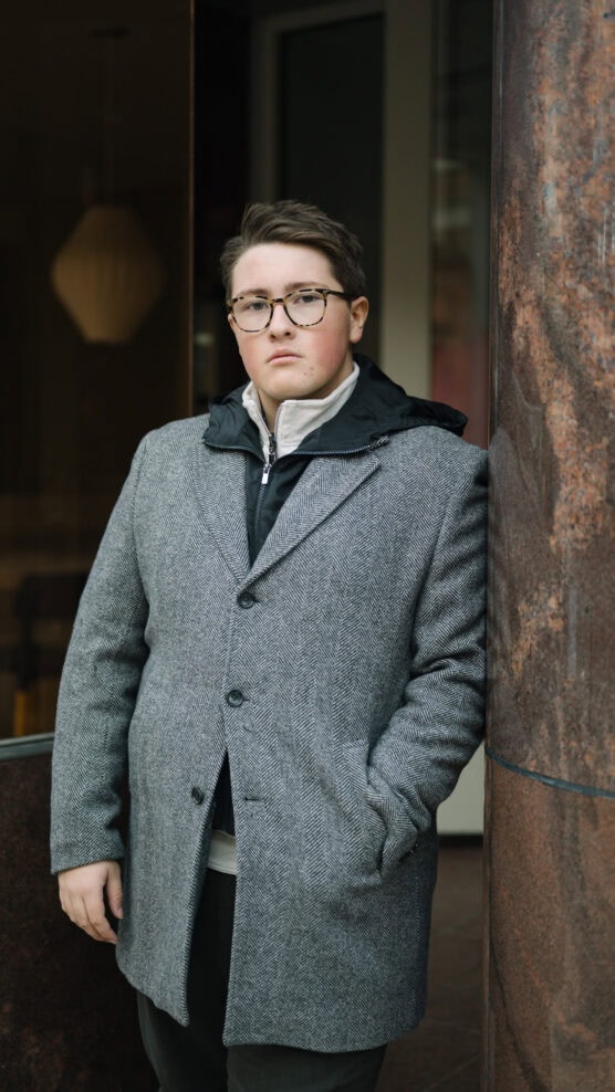 A person with short hair and glasses stands leaning against a marble column, wearing a grey coat over a zipped jacket, looking directly at the camera with a neutral expression. by Justin Salem Meyer