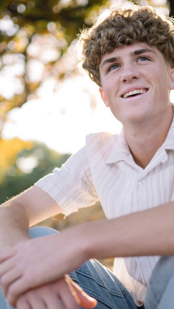 A young person with curly hair sits outdoors, smiling and looking slightly upward. They wear a light-colored, short-sleeve shirt and jeans, with trees and sunlight softly blurred in the background. by Justin Salem Meyer