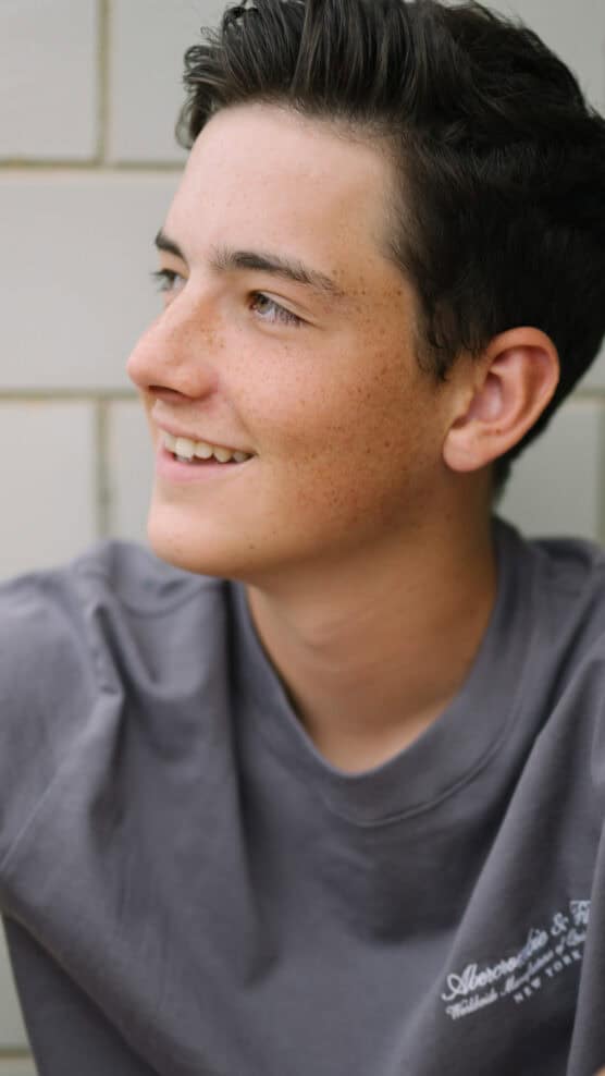 A teenage boy with short dark hair and freckles smiles and looks to the side. He is wearing a gray shirt and is in front of a tiled wall. by Justin Salem Meyer