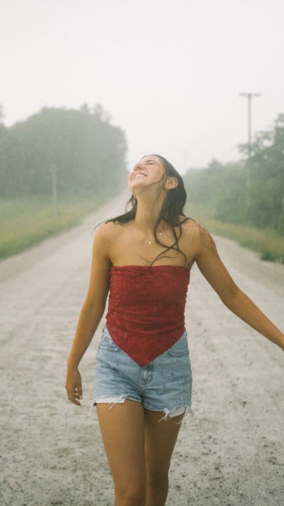 A young woman in a red strapless top and denim shorts stands on a gravel road, smiling and looking up as light rain falls, with greenery and telephone poles in the background. by Justin Salem Meyer