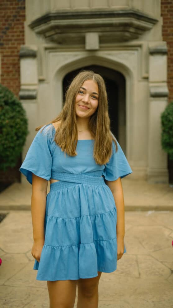 A teenage girl with long brown hair wearing a blue dress stands smiling outdoors in front of a stone building with an arched doorway and trimmed bushes on either side. by Justin Salem Meyer