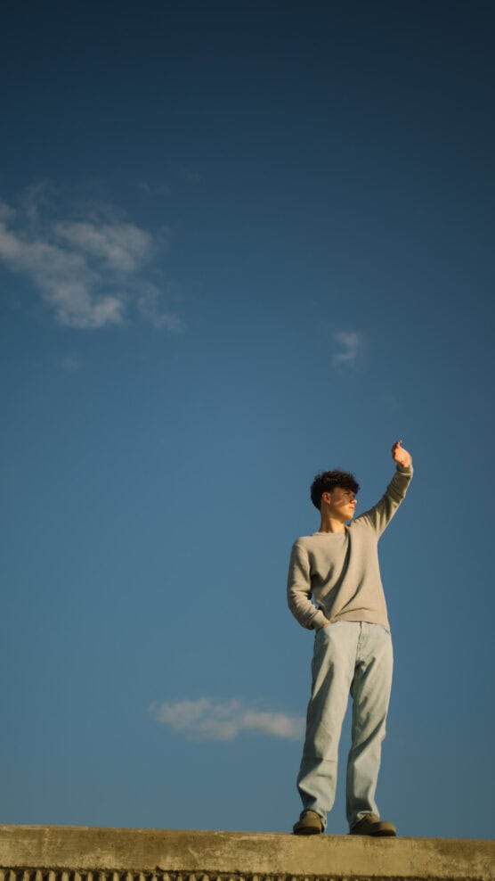 A person stands outdoors against a clear blue sky with a few clouds, wearing a gray sweater and light blue jeans, one hand in their pocket and the other raised while looking up. by Justin Salem Meyer