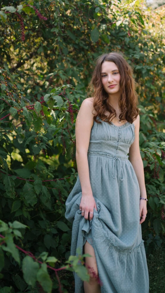 A young woman with wavy brown hair stands outdoors in front of leafy green bushes, wearing a light blue, sleeveless dress and gently holding up part of her skirt, looking calmly at the camera. by Justin Salem Meyer