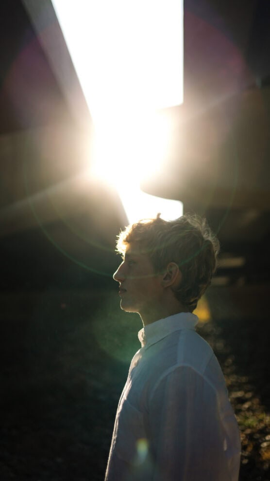 A person in a white shirt stands in profile under a structure with bright sunlight shining overhead, casting lens flares and creating a dramatic backlit effect. by Justin Salem Meyer