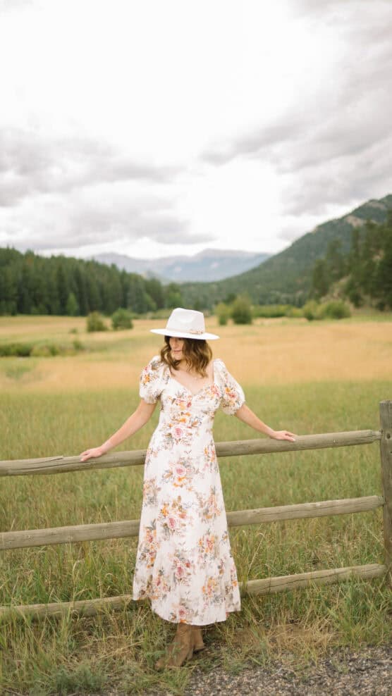 A woman in a white floral dress and white hat leans against a wooden fence in a grassy field with mountains and trees in the background under a cloudy sky. by Justin Salem Meyer