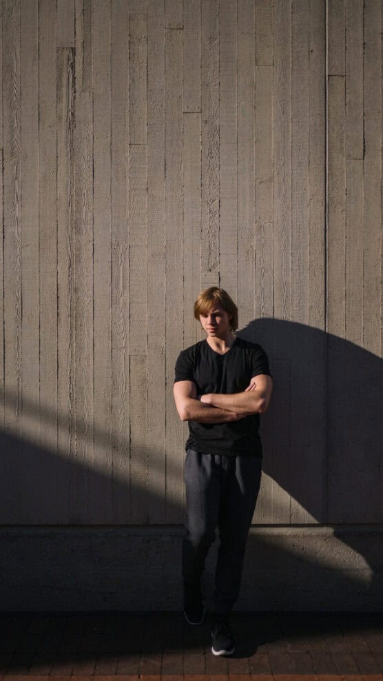 A young man with blond hair, wearing a black T-shirt and gray pants, stands with arms crossed against a tall concrete wall, partially lit by sunlight casting a shadow. by Justin Salem Meyer