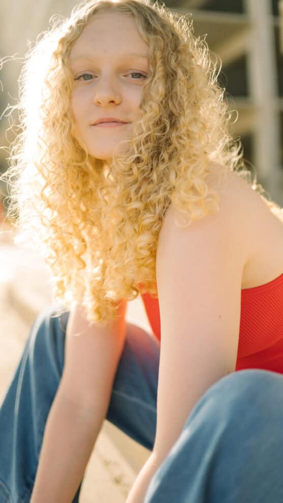 A person with long, curly blonde hair wearing a red sleeveless top and blue jeans sits outdoors in sunlight, looking confidently at the camera. by Justin Salem Meyer