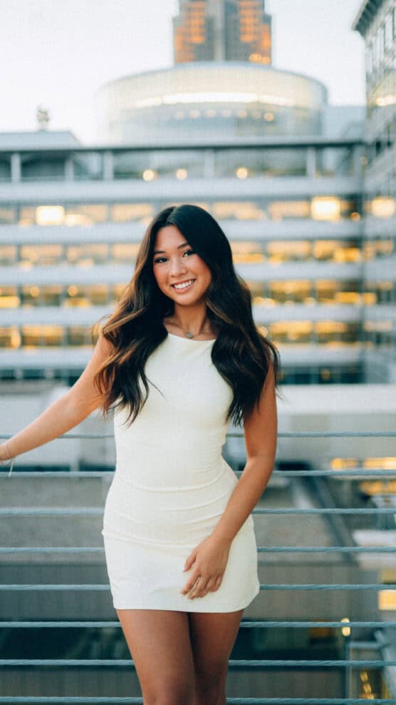 A young woman with long, wavy hair wearing a fitted white dress stands smiling on an outdoor balcony with a modern city building in the background. by Justin Salem Meyer