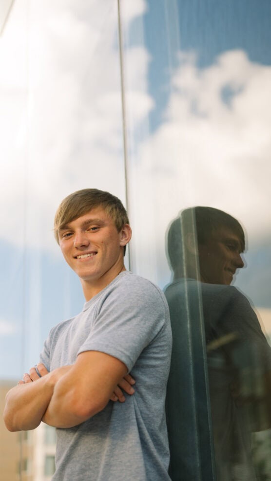 A young man with short light brown hair, wearing a gray t-shirt, stands with arms crossed and smiles, leaning against a reflective glass wall with clouds in the sky visible behind him. by Justin Salem Meyer