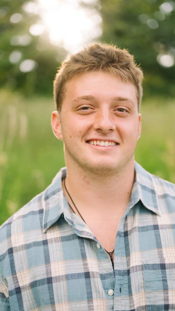 A young man with short, light brown hair smiles outdoors, wearing a blue plaid shirt. The background is blurred greenery with sunlight filtering through trees. by Justin Salem Meyer