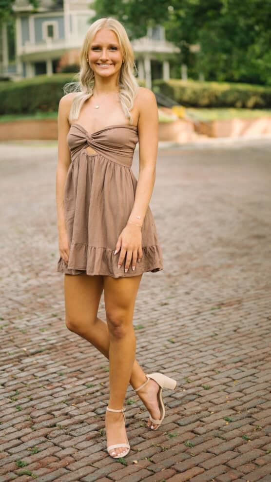 A young woman with long blonde hair stands outdoors on a cobblestone path, wearing a strapless brown dress and light-colored heeled sandals. She is smiling, with one leg slightly crossed in front of the other. Greenery and buildings are visible in the background. by Justin Salem Meyer