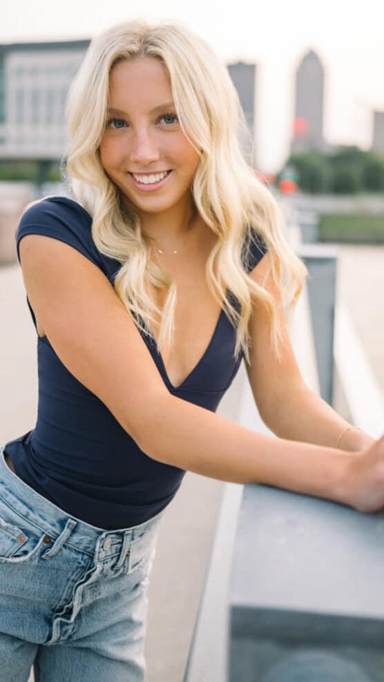 A young woman with long blonde hair, wearing a navy blue top and light blue jeans, leans on a railing outdoors with a cityscape blurred in the background. She is smiling at the camera. by Justin Salem Meyer