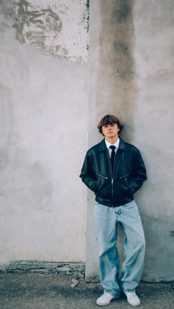 A young person with medium-length hair stands against a weathered, gray concrete wall, wearing a black leather jacket, white shirt, black tie, light blue baggy jeans, and white sneakers, with hands in their jacket pockets. by Justin Salem Meyer