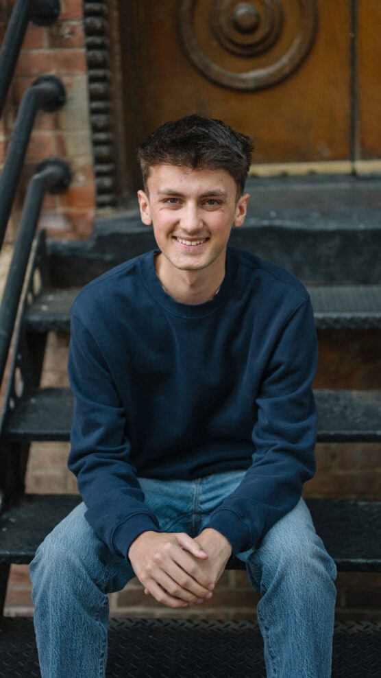 A young man with short brown hair, wearing a navy blue sweatshirt and light blue jeans, sits on outdoor black metal steps, smiling at the camera. A wooden door and brick wall are in the background. by Justin Salem Meyer