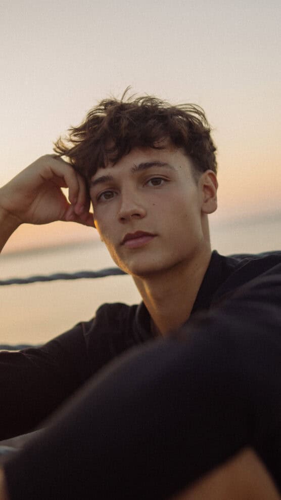 A young person with short, wavy brown hair sits outdoors at sunset, resting their head on one hand and looking calmly at the camera. The background shows a blurred horizon and soft, warm lighting. by Justin Salem Meyer