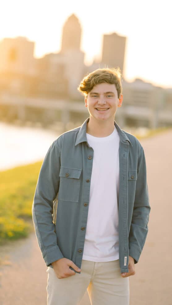 A young person with short, light brown hair smiles outdoors at sunset, wearing a white t-shirt and an open blue shirt-jacket, with a blurred city skyline in the background. by Justin Salem Meyer