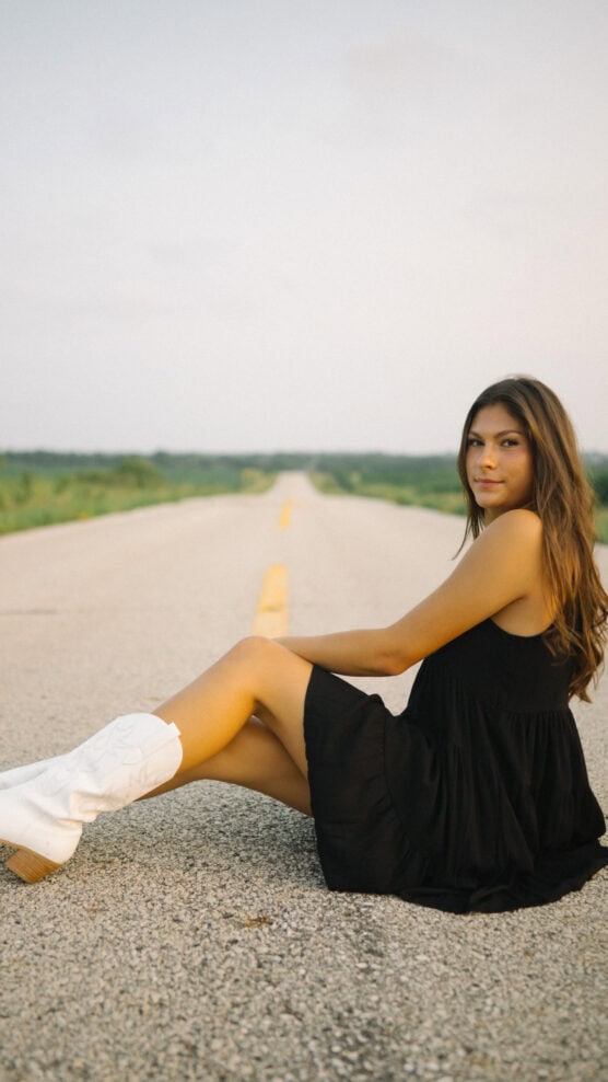 A young woman in a black dress and white cowboy boots sits on an empty rural road, looking at the camera. The background is green fields under a pale sky. by Justin Salem Meyer
