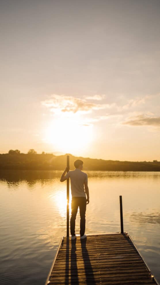 A person stands on a wooden dock by a calm lake at sunset, with golden sunlight reflecting on the water and a clear sky above. by Justin Salem Meyer