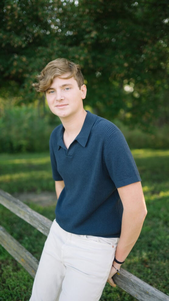 A young person with light brown hair, wearing a navy blue shirt and white pants, leans against a wooden fence outdoors with green trees and grass in the background. by Justin Salem Meyer