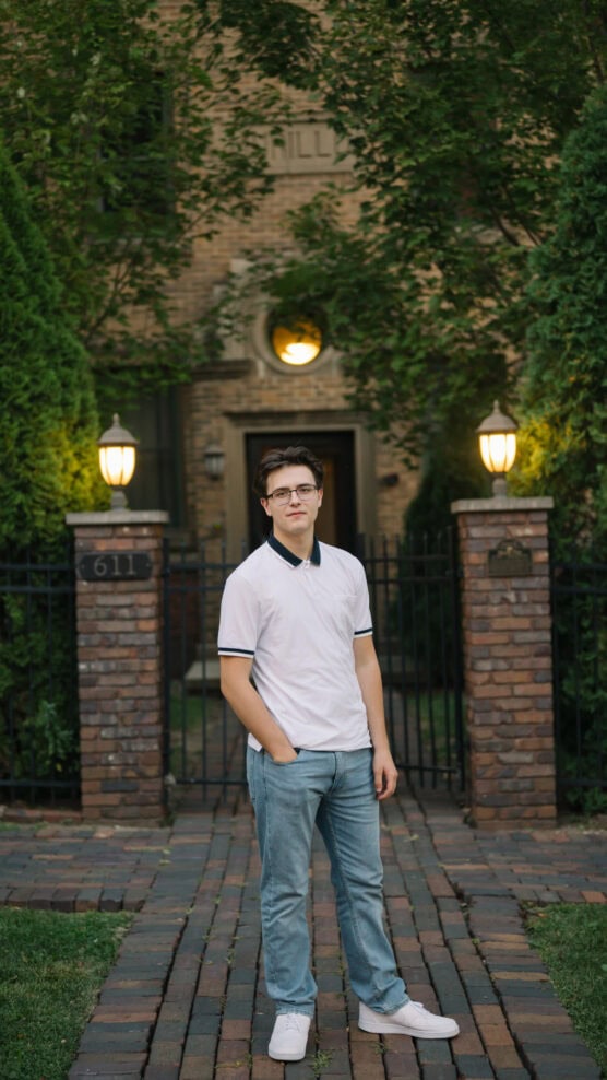 A young man wearing glasses, a white polo shirt, and jeans stands with one hand in his pocket on a brick pathway in front of a gated entrance to a brick building surrounded by trees and lit lamps. by Justin Salem Meyer