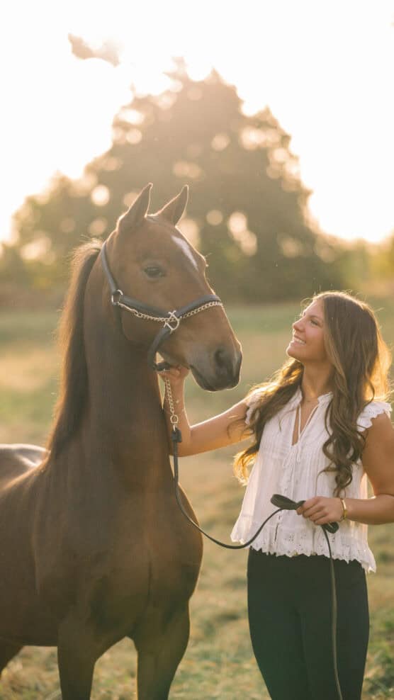 A young woman with long brown hair smiles at a brown horse while holding its reins in a sunlit field, with trees blurred in the background. by Justin Salem Meyer