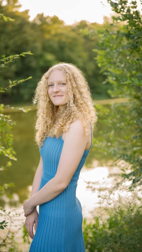 A young woman with curly blonde hair wearing a blue sleeveless dress stands outdoors near greenery and a calm body of water, smiling softly at the camera in natural sunlight. by Justin Salem Meyer