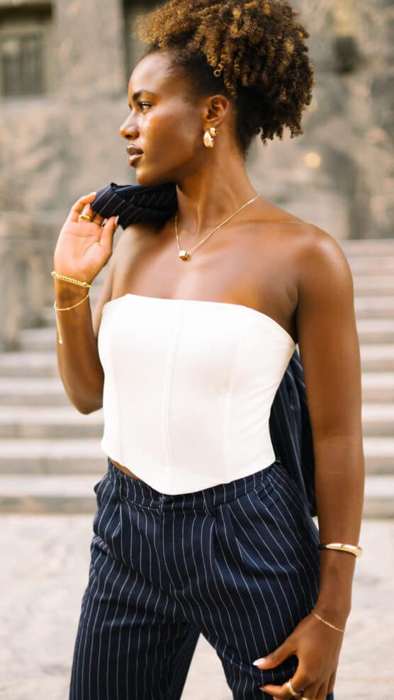 A woman stands outdoors, wearing a white strapless top and navy pinstripe pants, holding a matching pinstripe blazer over her shoulder. She has curly hair, gold jewelry, and looks confidently to the side. Steps are visible in the background. by Justin Salem Meyer