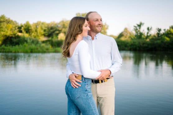 A couple stands close together by a calm lake, smiling and looking off into the distance. The woman wears a white top and jeans; the man wears a light shirt and khaki pants. Green trees and clear sky are in the background. by Justin Salem Meyer