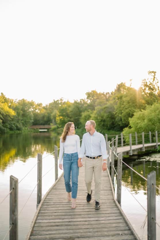 A couple walks hand-in-hand on a wooden footbridge over a calm lake, surrounded by lush green trees in soft sunlight. They are smiling at each other and dressed in casual, light-colored clothing. by Justin Salem Meyer