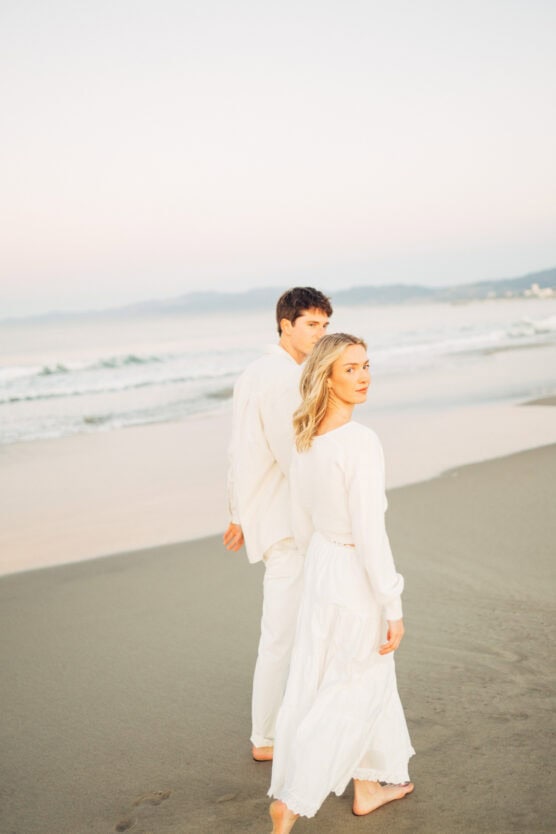 A man and woman dressed in white walk barefoot along a sandy beach at sunset, looking back towards the camera with waves and distant mountains in the background. by Justin Salem Meyer