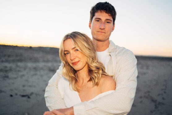 A couple stands close together on a sandy beach at sunset. The woman has blonde hair and smiles softly while the man stands behind her, arms wrapped around her shoulders, both looking at the camera. by Justin Salem Meyer