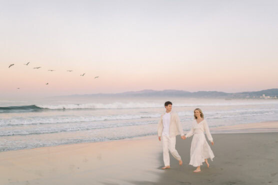A couple dressed in white walks barefoot along a beach at sunset, holding hands. Soft waves roll in, birds fly in the sky, and distant mountains are visible under a pastel-colored sky. by Justin Salem Meyer