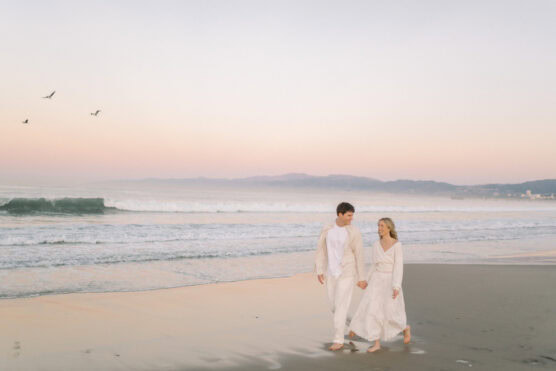 A couple dressed in white walks barefoot along a sandy beach at sunset, holding hands. Gentle waves roll in, distant mountains line the horizon, and four birds soar in the pastel-colored sky. by Justin Salem Meyer