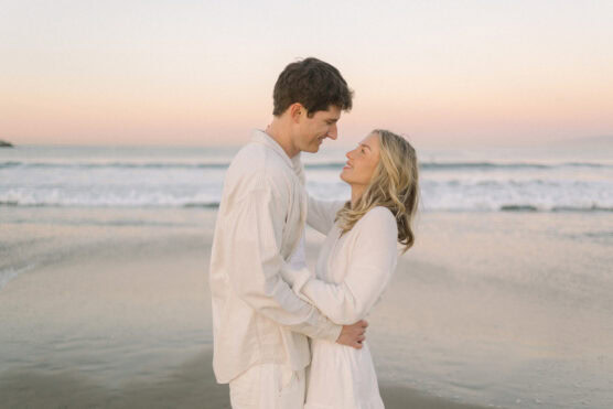 A couple dressed in white embraces and smiles at each other on a beach at sunset, with calm waves and a pastel-colored sky in the background. by Justin Salem Meyer