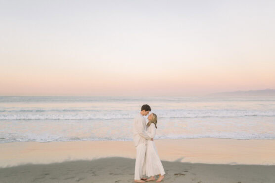 A couple dressed in white embraces and kisses on a sandy beach at sunset, with gentle ocean waves and a pastel sky in the background. by Justin Salem Meyer