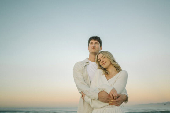 A couple stands close together at the beach during sunset. The woman leans her head on the mans chest as he wraps his arms around her, both looking peaceful. The sky is soft blue and pastel, with the ocean in the background. by Justin Salem Meyer