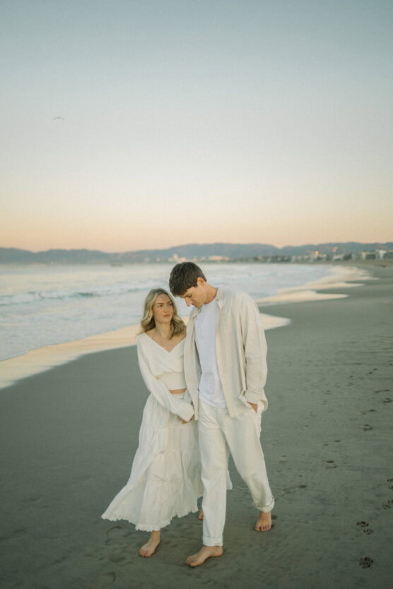 A young couple dressed in light, neutral clothing walks barefoot along a sandy beach at sunset, with gentle waves and distant mountains in the background. by Justin Salem Meyer