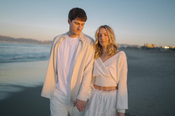A young couple wearing white clothes holds hands while walking on a beach at sunset, with soft sunlight highlighting their faces and the ocean in the background. by Justin Salem Meyer