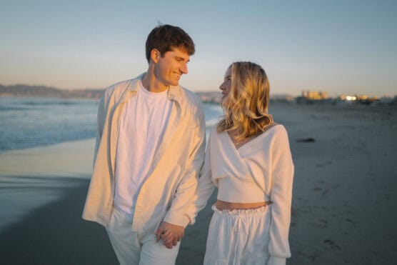 A young couple wearing light-colored clothes holds hands and smiles at each other while walking along a sandy beach at sunset. The ocean and distant buildings are visible in the background. by Justin Salem Meyer
