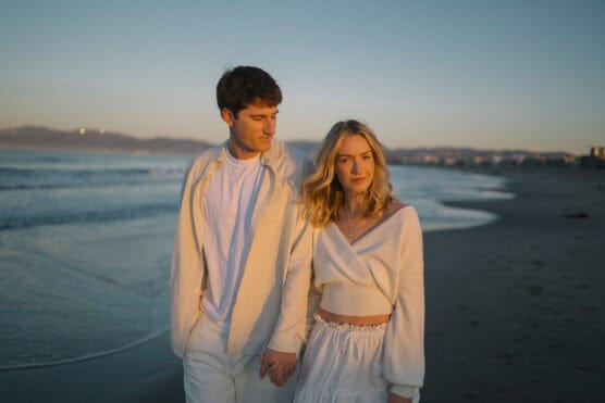 A young couple dressed in white holds hands and walks along a sandy beach at sunset, with gentle waves and distant hills in the background. by Justin Salem Meyer