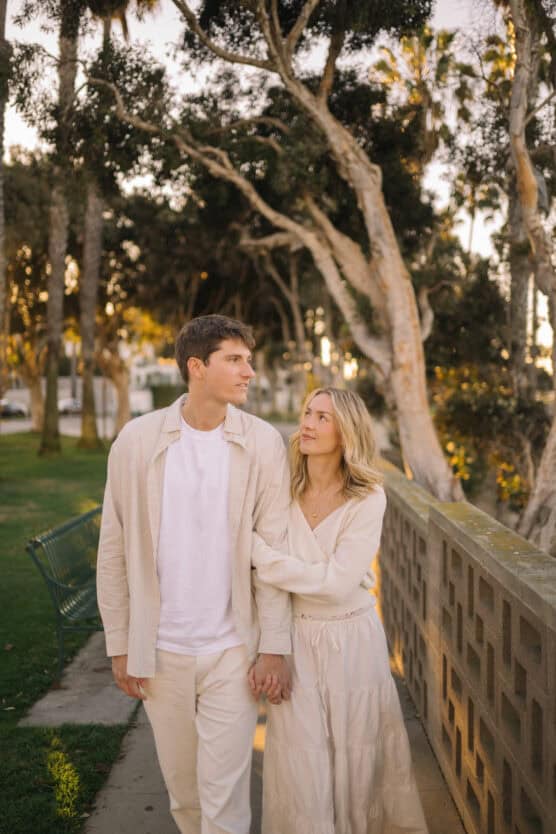A couple dressed in light, neutral outfits walks hand in hand along a sidewalk lined with trees. The woman looks up at the man, who gazes forward. They appear relaxed and content in the soft, golden sunlight. by Justin Salem Meyer