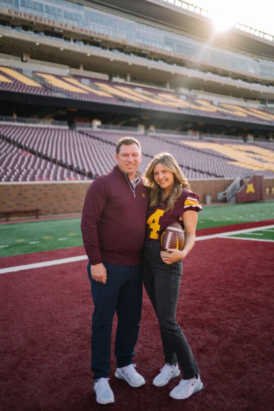 A man and woman stand on a football field in a stadium, smiling at the camera. The woman holds a football and wears a maroon jersey with the number 4. The stands and sunlight are visible in the background. by Justin Salem Meyer