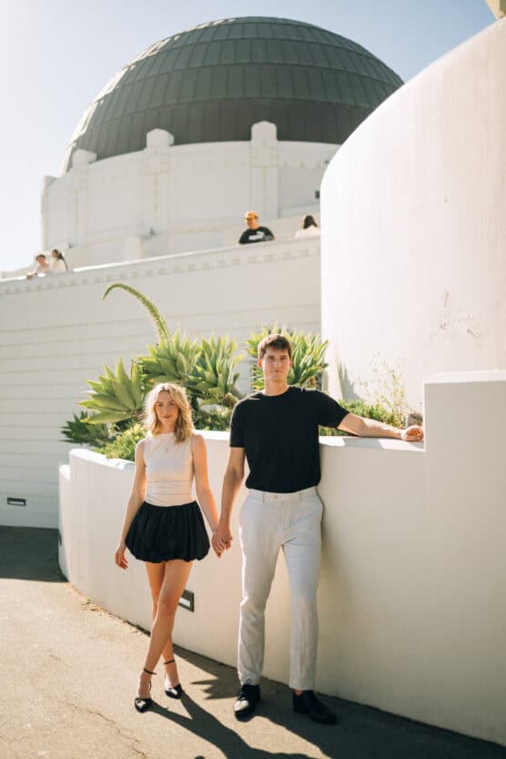 A young couple holds hands while standing outside in sunlight near white walls and greenery, with the Griffith Observatory’s domed roof visible in the background. by Justin Salem Meyer