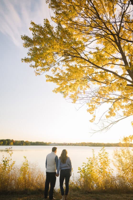 A couple stands hand in hand, facing a calm lake under a large tree with golden-yellow leaves, bathed in warm sunlight on an autumn day. by Justin Salem Meyer