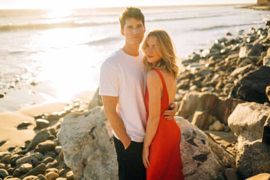 A couple stands on a rocky beach at sunset. The man wears a white t-shirt and black pants, while the woman wears a sleeveless red dress. They are close together, with the ocean and sunlight in the background. by Justin Salem Meyer