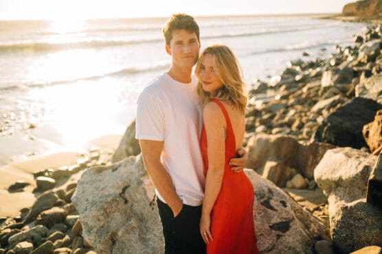 A couple stands on a rocky beach at sunset. The man wears a white t-shirt and black pants, while the woman wears a red dress. They embrace and look at the camera with the ocean and sunlight in the background. by Justin Salem Meyer